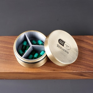 Peut inclure: Une boîte à pilules en laiton avec trois compartiments, remplie de capsules vertes. Le couvercle porte les inscriptions "Calm Down Cheer Up" et "Benjamin" gravées.