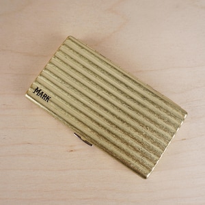 May include: A gold-colored, rectangular case with a ridged texture. The word "MARK" is printed in black on the top left corner. The case is lying on a light-colored wooden surface.