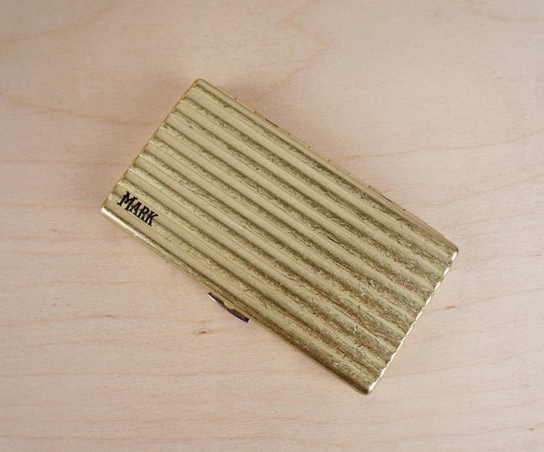 May include: A gold-colored, rectangular case with a ridged texture. The word "MARK" is printed in black on the top left corner. The case is lying on a light-colored wooden surface.
