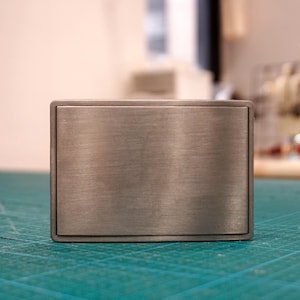May include: A rectangular belt buckle with a brushed silver-tone metal finish. The buckle features a smooth, flat front surface with a slightly raised border frame, presenting a clean and simple design with a metallic texture and subtle sheen.