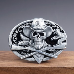 May include: Silver belt buckle with a skull wearing a cowboy hat and a snake. The skull has a bandana and two crossed rifles. The hat has the letter "R" on it.