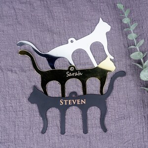 May include: Three stacked cat-shaped metal ornaments with personalized names. The top ornament is silver, the middle ornament is gold, and the bottom ornament is black. The names "Sarah" and "Steven" are engraved on the ornaments.