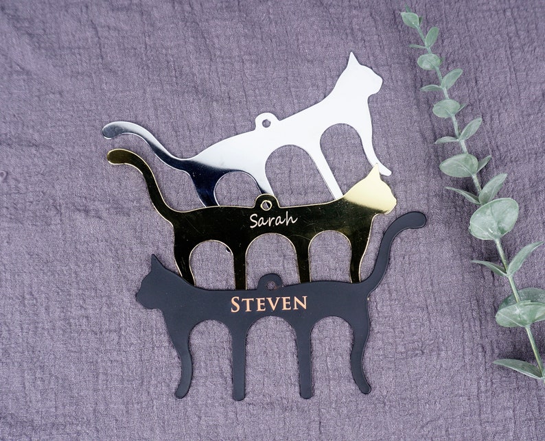 May include: Three stacked cat-shaped metal ornaments with personalized names. The top ornament is silver, the middle ornament is gold, and the bottom ornament is black. The names "Sarah" and "Steven" are engraved on the ornaments.