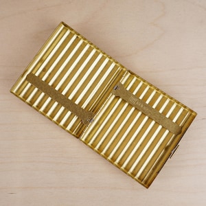 May include: Open, gold-colored metal cigarette case with vertical striped design. The case has a rectangular shape with a hinged lid and a clasp closure. Inside, there are two metal holders. The case is set against a light wooden surface.