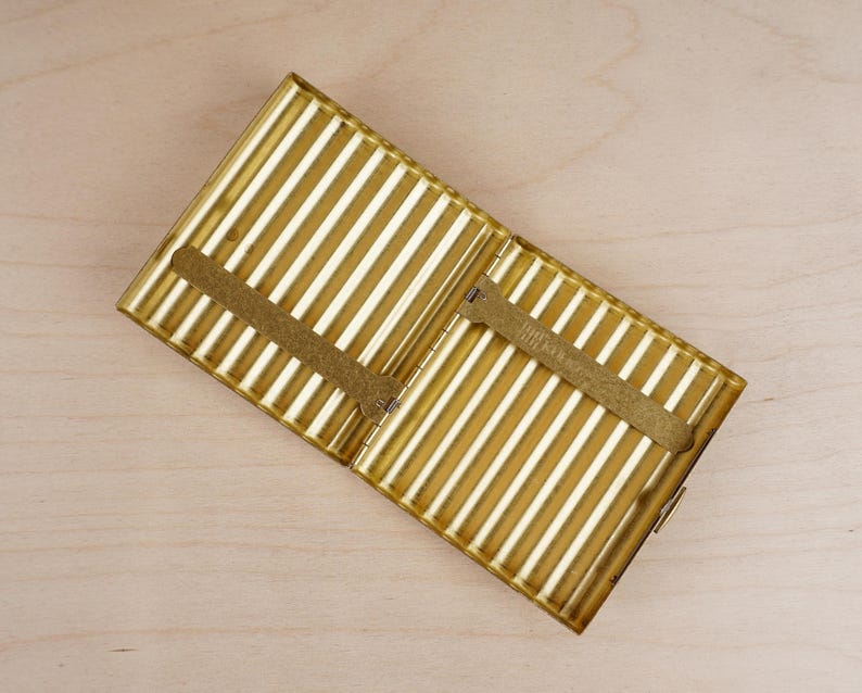 May include: Open, gold-colored metal cigarette case with vertical striped design. The case has a rectangular shape with a hinged lid and a clasp closure. Inside, there are two metal holders. The case is set against a light wooden surface.