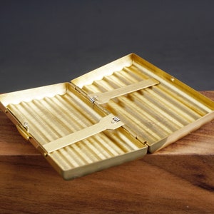 May include: Open, gold-colored cigarette case with a ribbed interior. The case is rectangular with rounded corners and a hinged lid. The case is open, revealing two metal holders. The case is on a wooden surface.