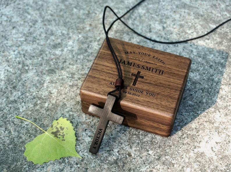 May include: A wooden box with a personalized inscription, "May your faith always guide you", and the name "JAMES SMITH" with a cross and the date "Sep 18, 2022". A wooden cross necklace with the word "VICTORY" engraved on it is resting on the box.