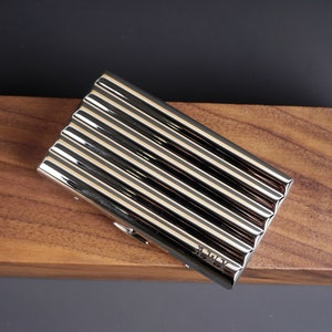 May include: A shiny, silver metal business card holder with a ribbed design. The rectangular case has a hinged opening and is set against a dark background and a wooden surface.
