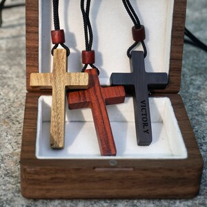 May include: Three wooden cross necklaces in a wooden box. The crosses are made of different types of wood: light brown, dark brown, and red. The dark brown cross has the name "VICTOR.Y" engraved on it.