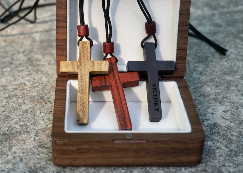 May include: Three wooden cross necklaces in a wooden box. The crosses are made of different types of wood: light brown, dark brown, and red. The dark brown cross has the name "VICTOR.Y" engraved on it.