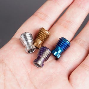 May include: Four small metal caps in different colours: silver, gold, blue, and purple. The caps have a threaded design and are arranged on a hand.