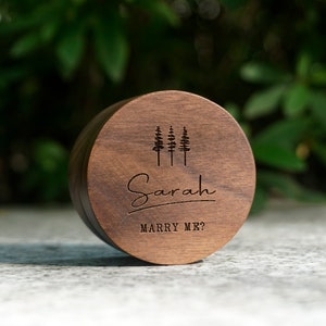 Puede incluir: Una caja de anillo de madera con un acabado marrón oscuro. La caja tiene un diseño de cuatro árboles y el texto "Sarah Marry Me?" grabado en la parte superior.
