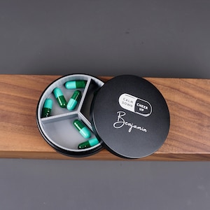 May include: Black pill organizer with three compartments, filled with green and white pills. The lid has the words "Calm Down" and "Cheer Up" printed on it, along with the name "Benjamin".