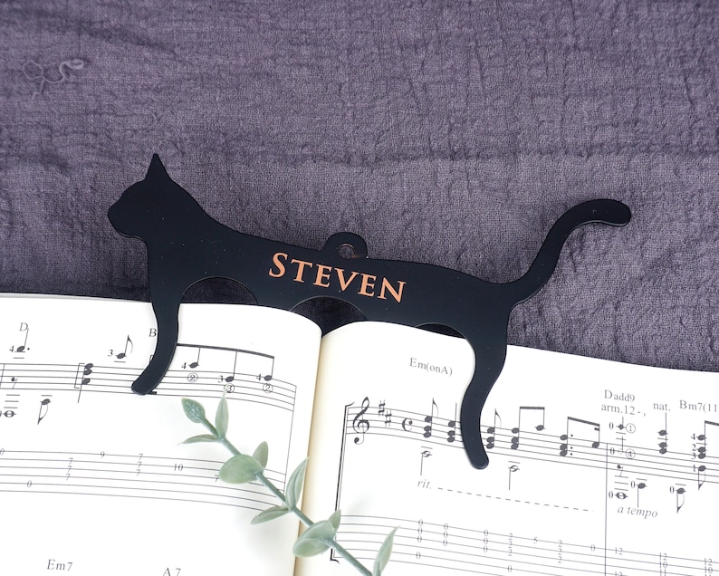 May include: Black metal cat bookmark with the name "STEVEN" printed in gold lettering. The bookmark is on top of a sheet of music.