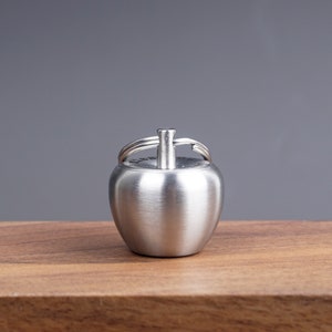 Puede incluir: Un llavero de plata con forma de manzana con un anillo en la parte superior.