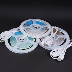 May include: Three spools of LED strip lights in blue, green, and white, each coiled on a clear plastic reel. White USB cables are attached. The background is black.