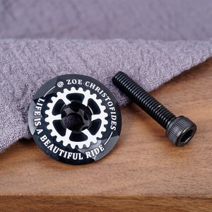 K&ouml;nnte beinhalten: Schwarzer Fahrradlenker-Deckel mit wei&szlig;er Beschriftung "@ZOE CHRISTOFIDES LIFE IS A BEAUTIFUL RIDE". Der Deckel liegt neben einer schwarzen Schraube.