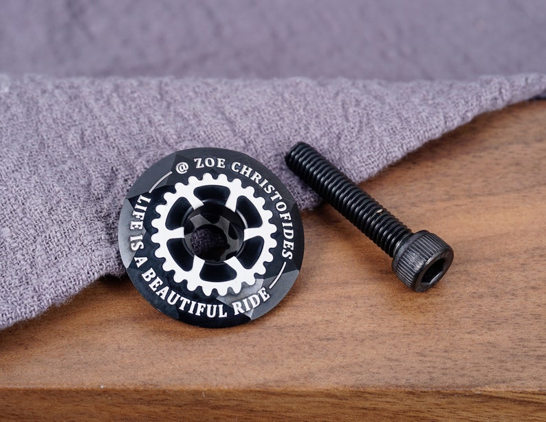 K&ouml;nnte beinhalten: Schwarzer Fahrradlenker-Deckel mit wei&szlig;er Beschriftung "@ZOE CHRISTOFIDES LIFE IS A BEAUTIFUL RIDE". Der Deckel liegt neben einer schwarzen Schraube.