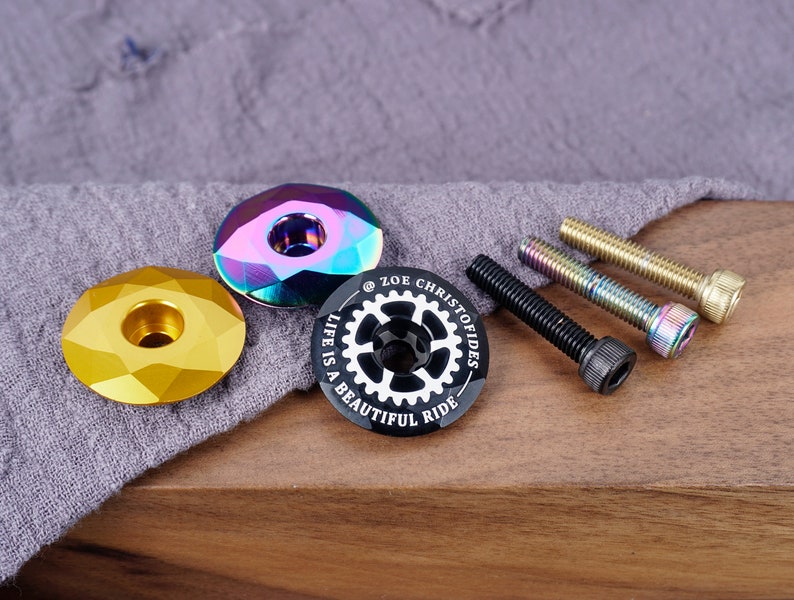 K&ouml;nnte beinhalten: Ein Satz aus drei Fahrradlenkerkappen, eine goldfarben, eine schillernd und eine schwarz mit dem Text "Life is a beautiful ride" und einem Zahnrad-Design. Das Set enth&auml;lt au&szlig;erdem drei Schrauben.