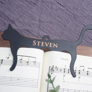 May include: Black metal cat-shaped bookmark with the name "STEVEN" engraved on it. The bookmark is holding open a book of sheet music.