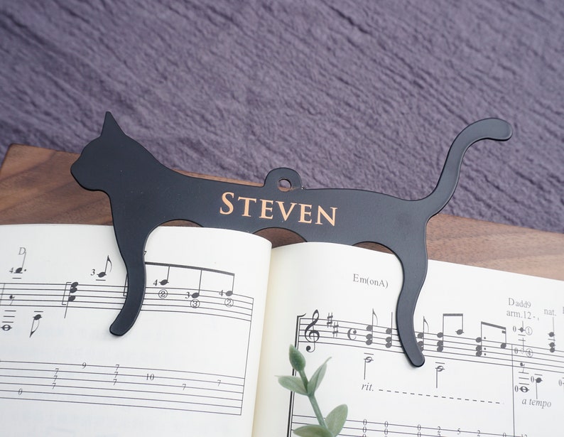 May include: Black metal cat-shaped bookmark with the name "STEVEN" engraved on it. The bookmark is holding open a book of sheet music.