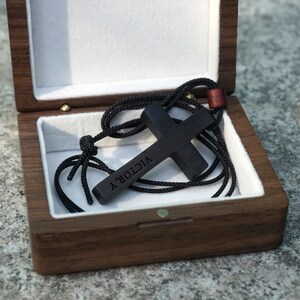 May include: A black wooden cross necklace with the word "VICTORY" engraved on it, inside a wooden box with a white velvet lining.