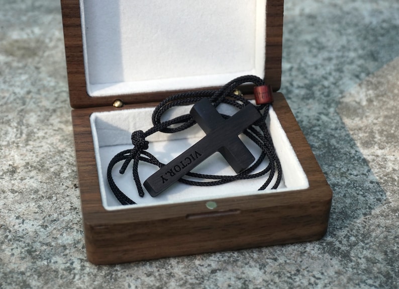 May include: A black wooden cross necklace with the word "VICTORY" engraved on it, inside a wooden box with a white velvet lining.