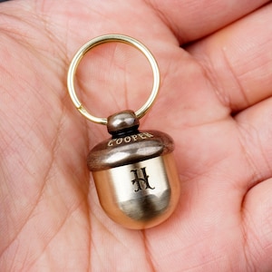 May include: A brass acorn keychain with a brown top and a gold bottom. The keychain has a ring and the word "COOPER" engraved on the top. The acorn has the letter "H" engraved on the front.
