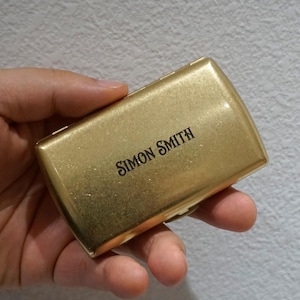 May include: A gold-colored rectangular box with rounded edges, held in a hand. The text "SIMON SMITH" is printed in black on the top of the box. The box has a shiny, metallic finish.