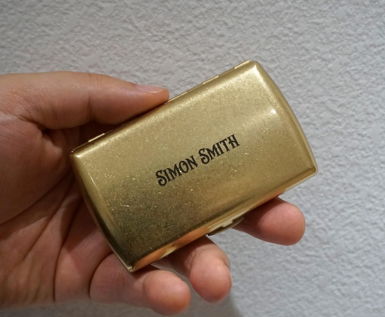 May include: A gold-colored rectangular box with rounded edges, held in a hand. The text "SIMON SMITH" is printed in black on the top of the box. The box has a shiny, metallic finish.