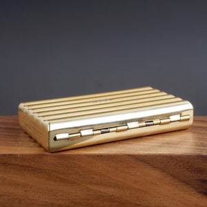 May include: A rectangular, gold-colored metal box with a ribbed design on the top. The box has a hinged lid and is resting on a wooden surface. The box appears to be a vintage or antique item.