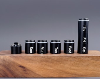 Peut inclure: Un pilulier hebdomadaire noir avec sept compartiments cylindriques. Chaque compartiment est étiqueté avec un jour de la semaine en texte blanc. Le pilulier est en métal et possède un couvercle à visser. Le pilulier est sur une surface en bois.