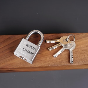 May include: A silver padlock with the text "SUS304/NICKEL 40mm" and "RAPHAEL COLEMAN" engraved on the front. The padlock is accompanied by three silver keys.