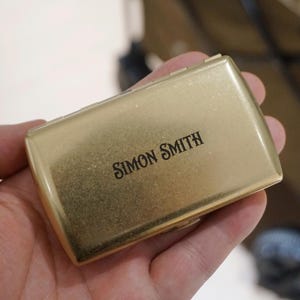 May include: A gold-colored rectangular case with rounded edges, held in a hand. The case has the words "SIMON SMITH" printed in black, centered on the top. The case appears to be made of metal.