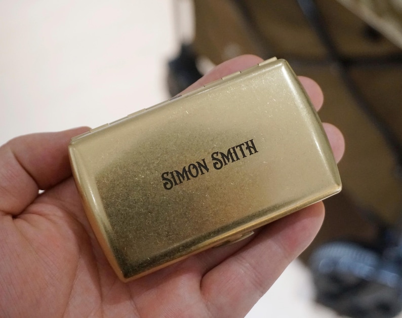 May include: A gold-colored rectangular case with rounded edges, held in a hand. The case has the words "SIMON SMITH" printed in black, centered on the top. The case appears to be made of metal.