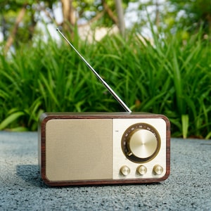 Pu&ograve; includere: Una radio in legno in stile vintage con quadrante e manopole dorate. La radio ha un'antenna argentata ed &egrave; appoggiata su una superficie grigia.