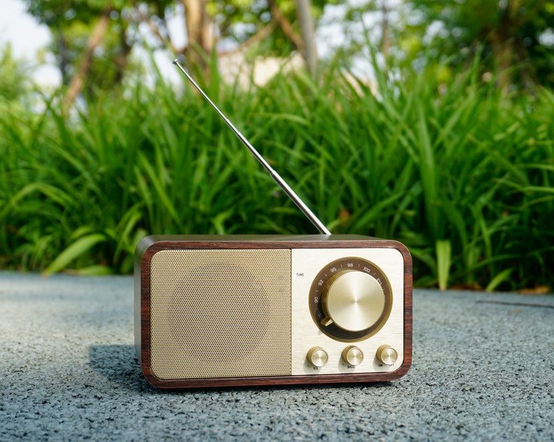 Pu&ograve; includere: Una radio in legno in stile vintage con quadrante e manopole dorate. La radio ha un'antenna argentata ed &egrave; appoggiata su una superficie grigia.