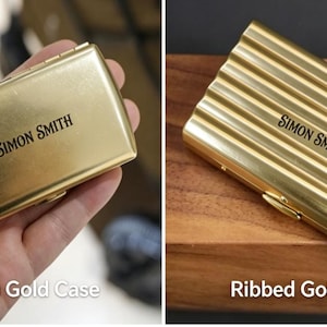 May include: Gold-colored business card case with the name "SIMON SMITH" printed in black. The case is rectangular and has a smooth surface. The image also shows a ribbed gold case with the same name.