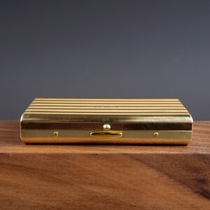 May include: A rectangular, gold-colored case with horizontal lines. The case has a clasp and small rivets. The object is on a wooden surface with a dark background. The case appears to be made of metal.