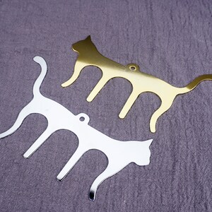 May include: Two metal cat-shaped ornaments, one gold and one silver, with a cutout design. The ornaments are on a textured gray surface.