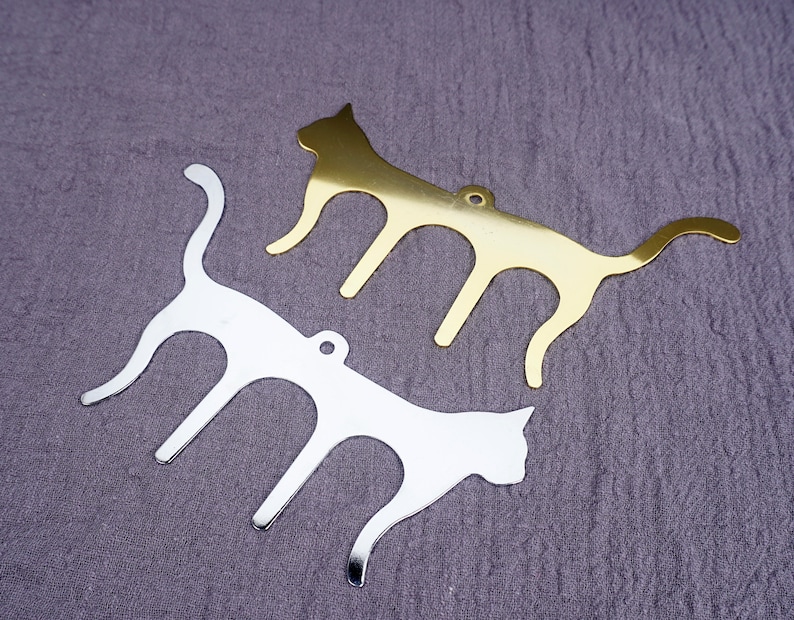May include: Two metal cat-shaped ornaments, one gold and one silver, with a cutout design. The ornaments are on a textured gray surface.