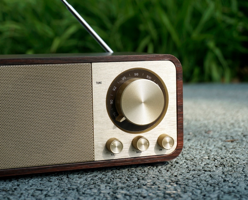 Pu&ograve; includere: Una radio in legno in stile vintage con un quadrante e manopole dorate. La radio ha una griglia dell'altoparlante con un motivo a maglia fine. Il quadrante ha numeri da 92 a 100 e le parole "TUNE", "BASS" e "VOL".