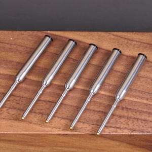 May include: Five silver-toned metal pen refills arranged in a parallel row. Each refill features a cylindrical body, a tapered tip, and a small gold-colored ballpoint at the end. The top of each refill is capped with a black circular plug.