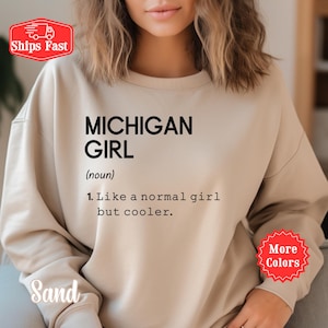Könnte beinhalten: Ein sandfarbenes Sweatshirt mit Rundhalsausschnitt und schwarzem Textaufdruck: MICHIGAN GIRL (noun) 1. Like a normal girl but cooler. Das Kleidungsstück verfügt über lange Ärmel und einen gerippten Halsausschnitt in einem schlichten Design.