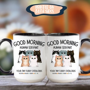 Puede incluir: Taza de caf&eacute; de cer&aacute;mica blanca con borde y asa negros. La taza tiene un gr&aacute;fico de cinco gatos de dibujos animados con el texto "Good Morning Human Servant" y "Your Tiny Furry Overlords" con los nombres "Trooper, Peaches, Raisin, ET, Al".