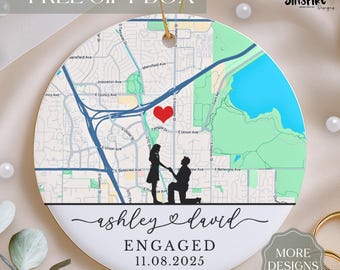 May include: A round white ceramic ornament featuring a printed street map with a red heart icon, a black silhouette of a couple, and the text "ashley & david", "ENGAGED", and "11.08.2025". It includes a gold hanging string at the top center.