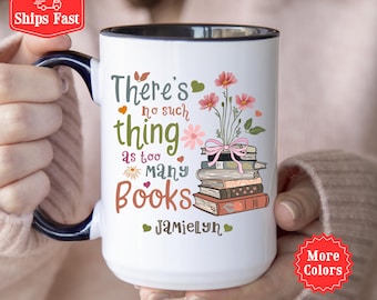 May include: White ceramic mug with a black interior and handle. The mug features the text "There's no such thing as too many books" with floral and heart accents. A stack of books and flowers are also displayed.