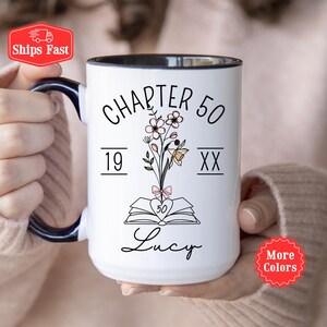Peut inclure: Un mug en céramique blanche avec une anse et un bord intérieur bleu foncé. Le motif présente le texte "CHAPTER 50" au-dessus d'une illustration de fleurs sortant d'un livre ouvert. Les chiffres "19" et "XX" entourent les fleurs, avec "Lucy" en dessous.