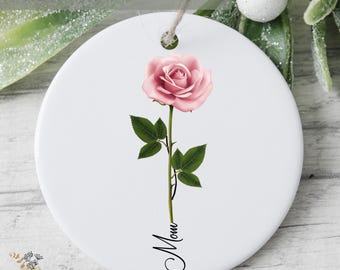 May include: A round white ceramic ornament featuring a single pink rose with green leaves on a stem. The word Mom is written in black cursive script extending from the bottom of the stem. A small hole at the top allows for hanging.