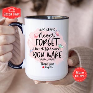 May include: White ceramic coffee mug with a black rim and handle. The mug features a pink watercolor background with butterflies and the text "Mrs. Gravis Never Forget the difference YOU MAKE Thank you! Jayda".